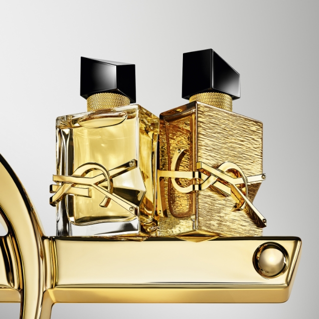 Due squisite bottiglie di profumo YSL Libre, una trasparente e una dorata testurizzata, sono esposte fianco a fianco su un lussuoso elemento scultoreo dorato