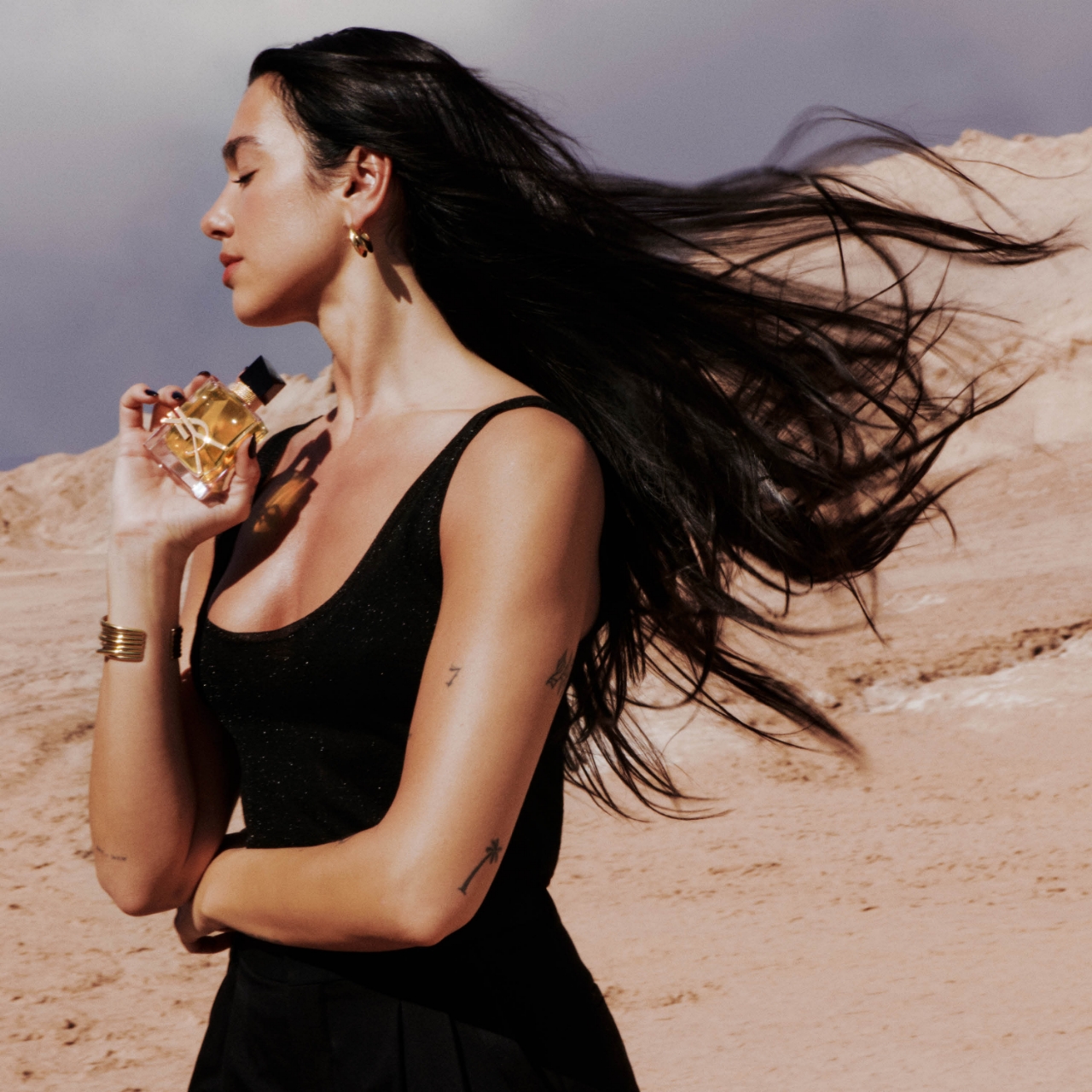 Dua Lipa, con i capelli che volano al vento, tiene la bottiglia del profumo YSL Libre su uno sfondo desertico caldo, con gli occhi chiusi in serenità