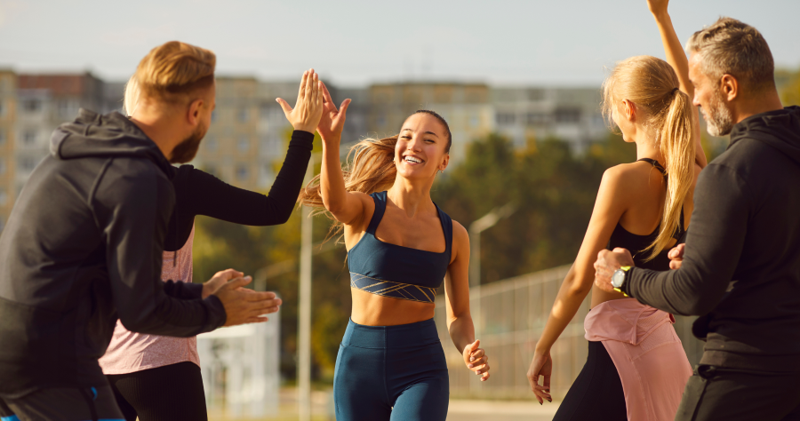 alt="Gruppo di adulti in forma all'aperto che festeggiano con il cinque dopo un allenamento o una corsa, sorridenti ed energizzati sotto il sole.»
