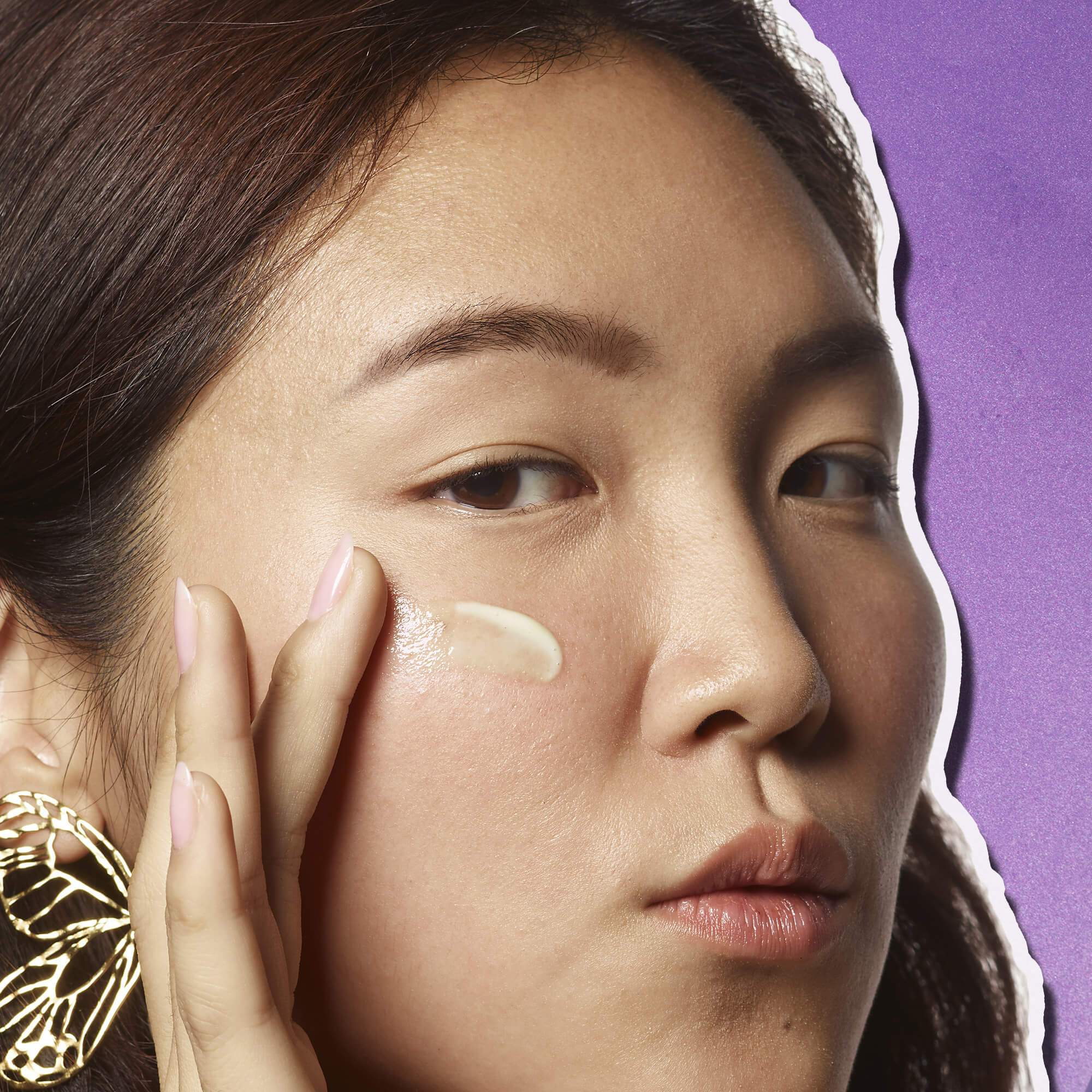 Donna con orecchino dorato a farfalla che applica il siero Kiehl’s sulla guancia, su sfondo viola.