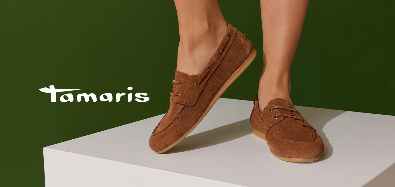 Scarpini slip-on in camoscio marrone con punta tonda, dettagli in laccio e suola in gomma chiara, esposti su un pedestal bianco.