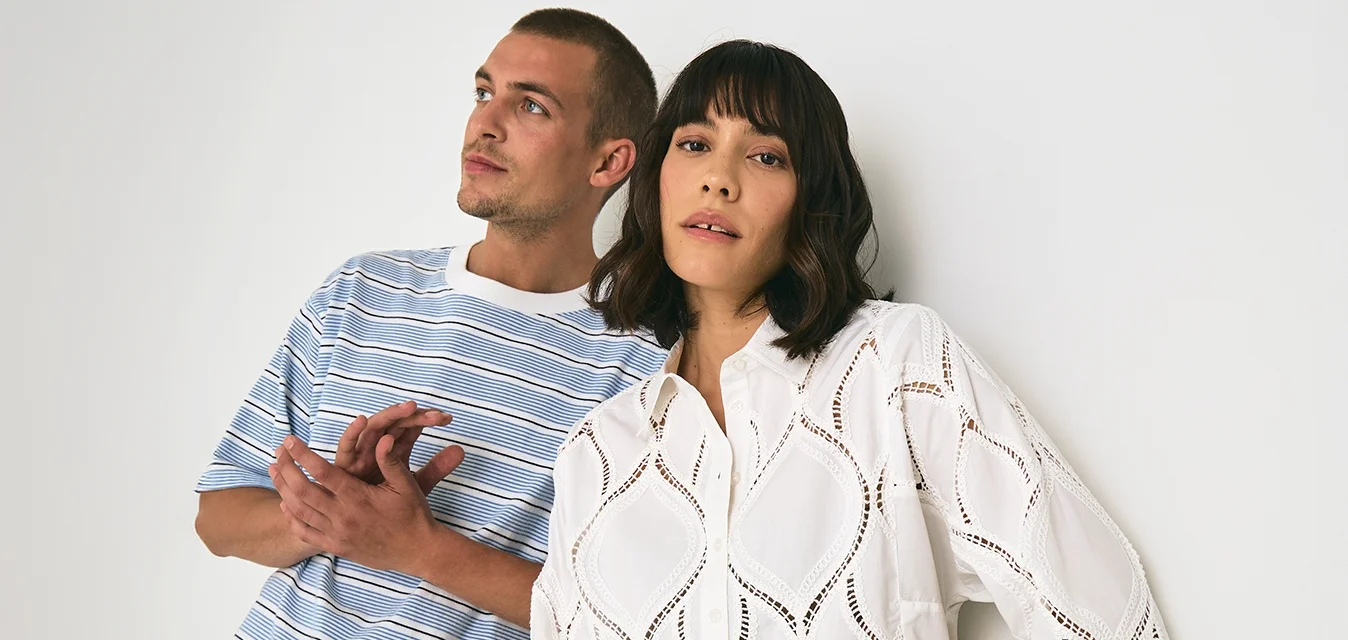 Maglietta a righe blu e bianca abbinata a pantaloni beige, insieme a una camicia di pizzo bianca con ritagli a diamante e pantaloni a righe. Sfondo puro.