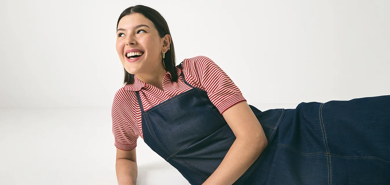 Abito a grembiule in denim con taglio lungo, abbinato a una maglietta a righe rosse e bianche. Il tessuto è liscio con dettagli di cucitura visibili.