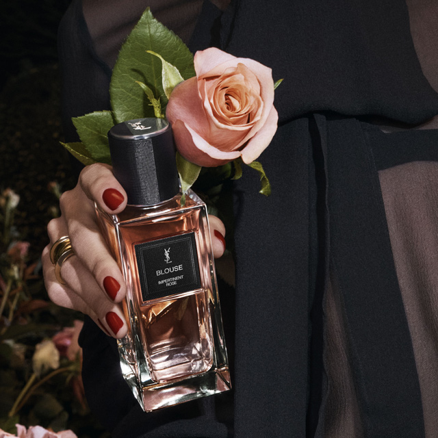 Una mano con unghie rosse smaltate e anelli d'oro tiene una bottiglia rettangolare del profumo YSL "BLOUSE IMPERTINENT ROSE". Una rosa rosa tenue con foglie verdi è artisticamente posizionata dietro la bottiglia, completando il colore delicato del profumo.