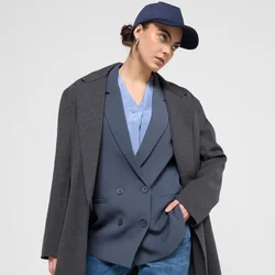 Blazer grigio oversize su un gilet blu doppiopetto, abbinato a pantaloni cargo blu. Il modello indossa un berretto blu navy, con uno sfondo a texture liscia.