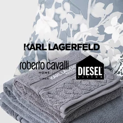 Pila di asciugamani grigi Karl Lagerfeld su una superficie bianca con cuscini a stampa floreale e leopardata sullo sfondo.