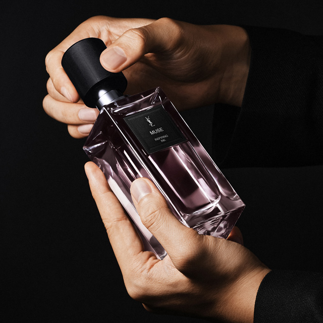Un paio di mani, probabilmente maschili, tengono delicatamente una bottiglia rettangolare del profumo YSL "MUSE IMPERTINENT ROSE". Le mani regolano il tappo nero della bottiglia dalla profonda tonalità viola, su uno sfondo nero intenso.