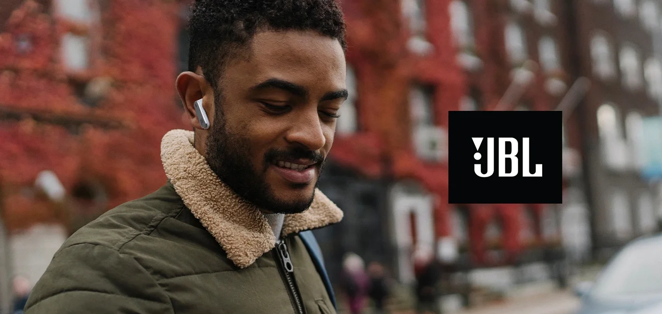 Uomo con cuffie e giacca verde guarda lo smartphone su una strada di città con edifici dalle foglie rosse e un'auto parcheggiata sullo sfondo.