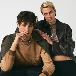 Giovane uomo seduto su uno sgabello, vestito con un maglione marrone e pantaloni neri, donna in piedi accanto a lui con giacca di pelle nera e gonna di pizzo su sfondo bianco.