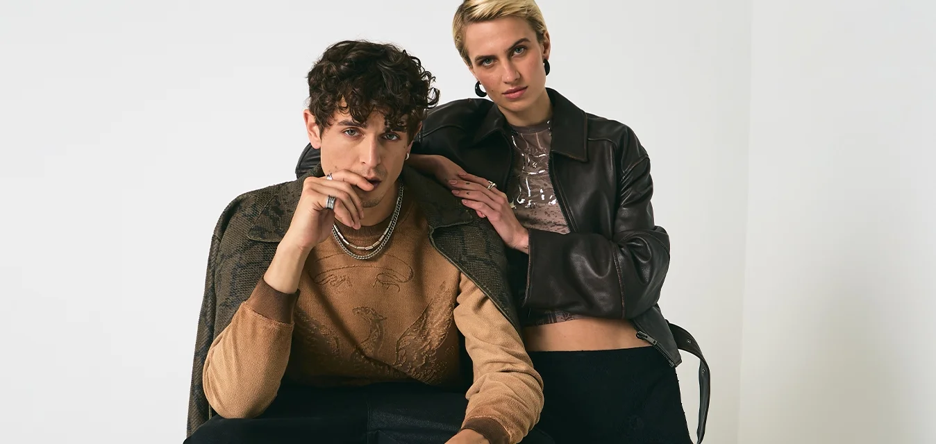 Giovane uomo seduto su uno sgabello, vestito con un maglione marrone e pantaloni neri, donna in piedi accanto a lui con giacca di pelle nera e gonna di pizzo su sfondo bianco.