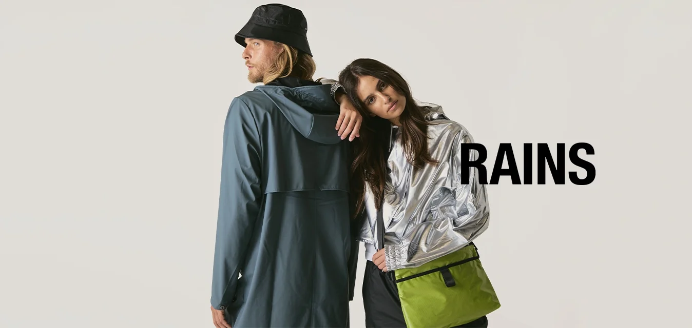 Un uomo con un impermeabile blu e un cappello a secchiello nero è in piedi schiena contro schiena con una donna in giacca argento che inclina la testa sulla sua spalla, mentre porta una borsa verde.