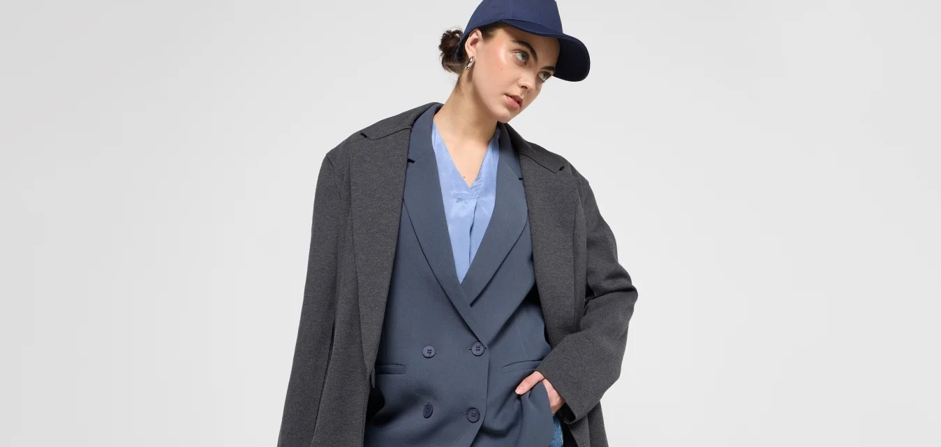 Blazer grigio oversize su un gilet blu doppiopetto, abbinato a pantaloni cargo blu. Il modello indossa un berretto blu navy, con uno sfondo a texture liscia.