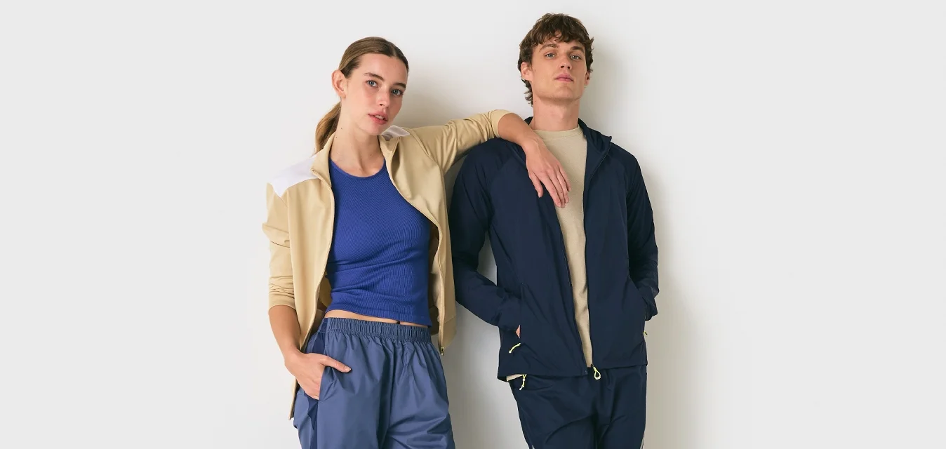 Due giovani adulti in abbigliamento sportivo casual appoggiati a un muro bianco, uno con il braccio appoggiato sulla spalla dell'altro.