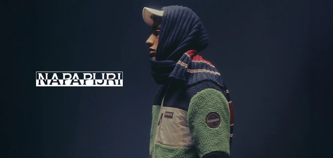 Persona di profilo che indossa una giacca di pile verde e nera, un cappello bianco e una sciarpa blu con strisce rosse e bianche su uno sfondo scuro.