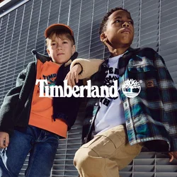 Due bambini sono in piedi contro un muro grigio con persiane. Uno indossa una giacca nera sopra una felpa arancione e jeans blu, con stivali neri. L'altro ha una giacca a quadri sopra una camicia bianca, pantaloni color khaki e scarpe gialle.