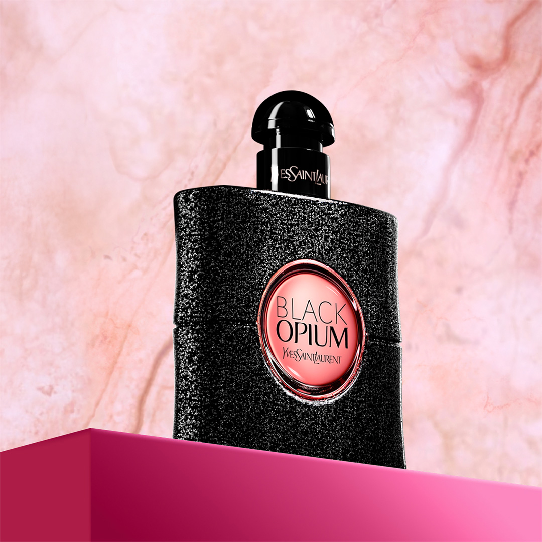 Un flacone di profumo Yves Saint Laurent Black Opium nero e scintillante con un'etichetta rosa è posato su una piattaforma fucsia su uno sfondo rosa tenue
