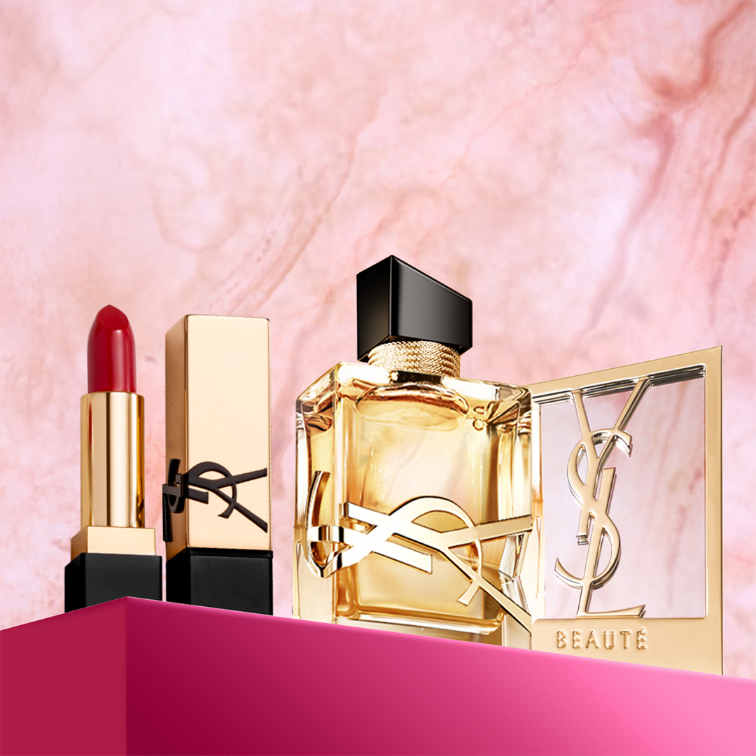 Un rossetto rosso Yves Saint Laurent, un flacone dorato del profumo YSL Libre e uno specchio compatto dorato con il logo YSL e "BEAUTÉ", il tutto esposto su una superficie rosa fucsia su uno sfondo di marmo rosa