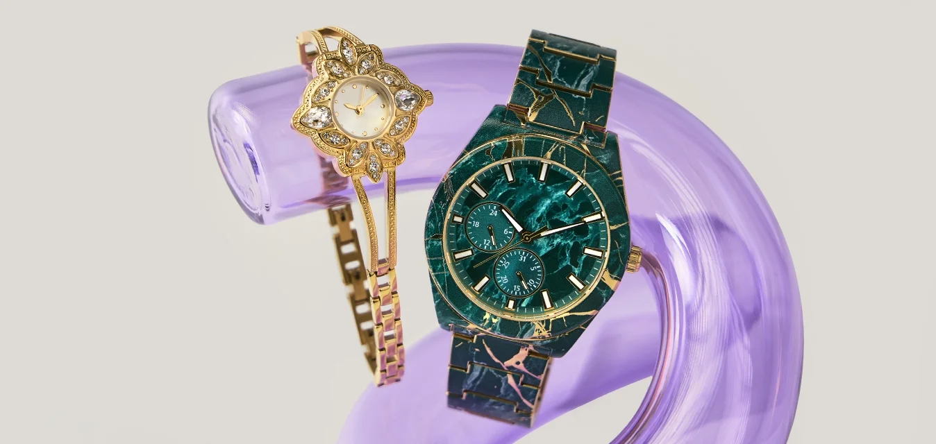 Orologio bracciale floreale dorato e orologio da polso con motivo marmo verde scuro poggiati su un supporto curvo traslucido viola su uno sfondo chiaro.