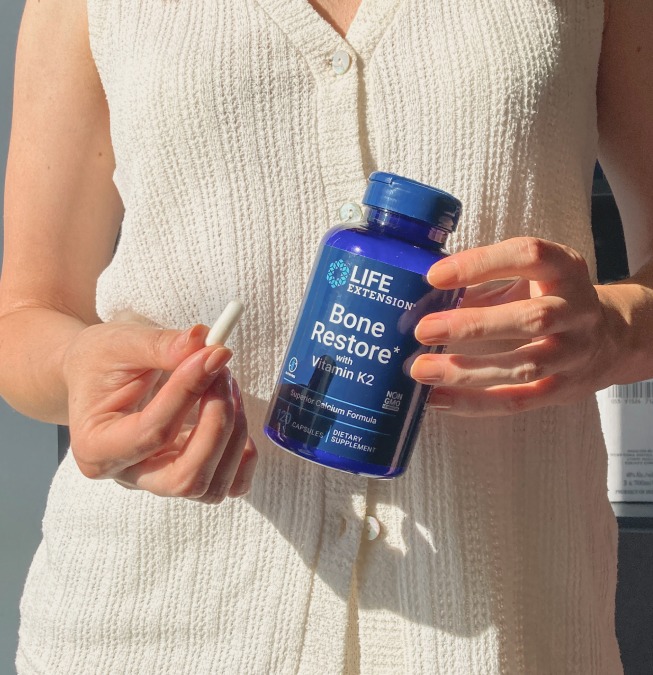 alt="Persona con un top in maglia bianca con in mano un flacone Life Extension Bone Restore con vitamina K2 e una capsula esposta alla luce naturale, che simboleggia la forza ossea e il sostegno quotidiano.»