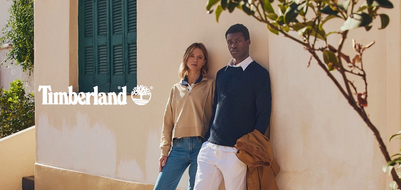 Due persone stanno su un terrazzo di pietra accanto a un muro giallo con persiane verdi, indossando abiti casual dai colori tenui.