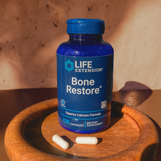 alt="Flacone di integratore Life Extension Bone Restore posto su un vassoio rotondo di legno con delle capsule accanto, alla calda luce naturale del sole.»
