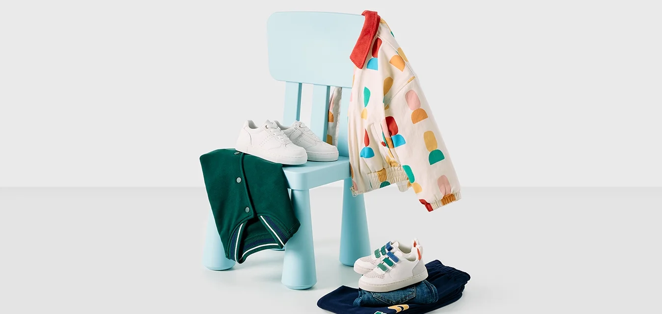 Sedia in legno azzurro con schienale arrotondato, una giacca rossa e colorata appoggiata sopra, e scarpe bianche su un indumento piegato di colore blu navy.