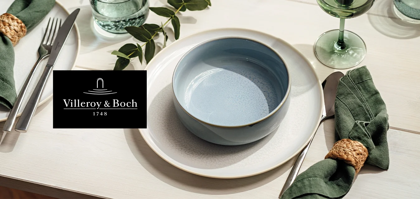 Piatti da pranzo bianchi con una ciotola blu, tovaglioli verdi in anelli intrecciati, posate, bicchieri trasparenti e verdi, e decorazione da tavolo con foglie.