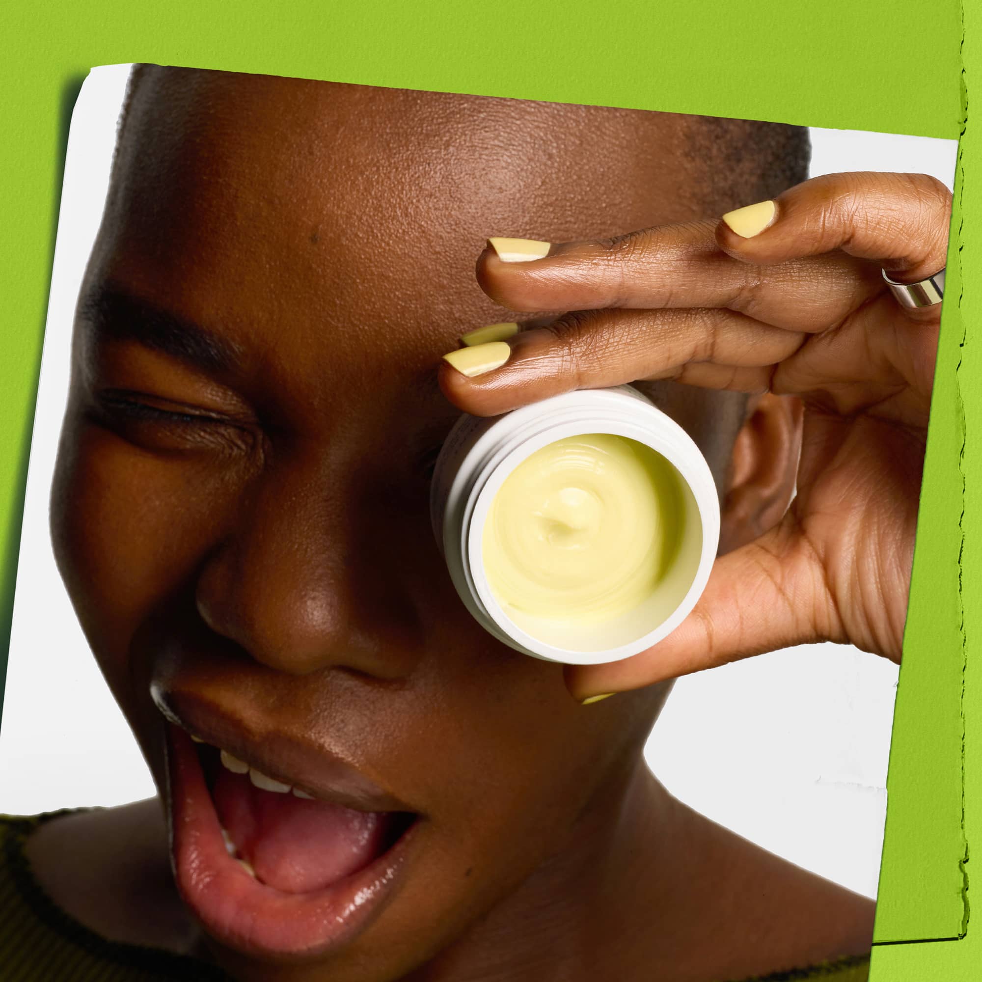 Persona che sorride con un occhio chiuso e tiene il vasetto di Creamy Eye Treatment with Avocado davanti all’altro occhio. Smalto e crema di colore giallo chiaro su sfondo verde.
