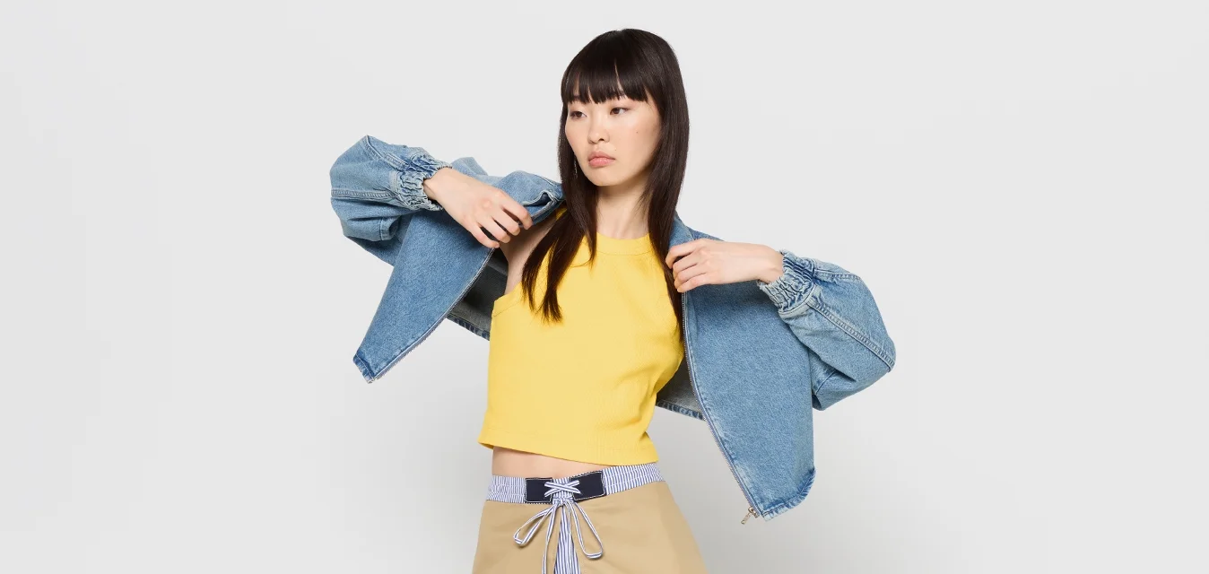 Giacca di denim con maniche a sbuffo, top giallo corto e gonna beige con una cravatta a righe. Texture morbide; contrasto di colori vivaci.