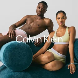 Uomo in boxer rosa che si sdraia sul bracciolo di un divano blu arrotondato, donna in reggiseno sportivo giallo a coste e pantaloni corti che si siede accanto a lui.