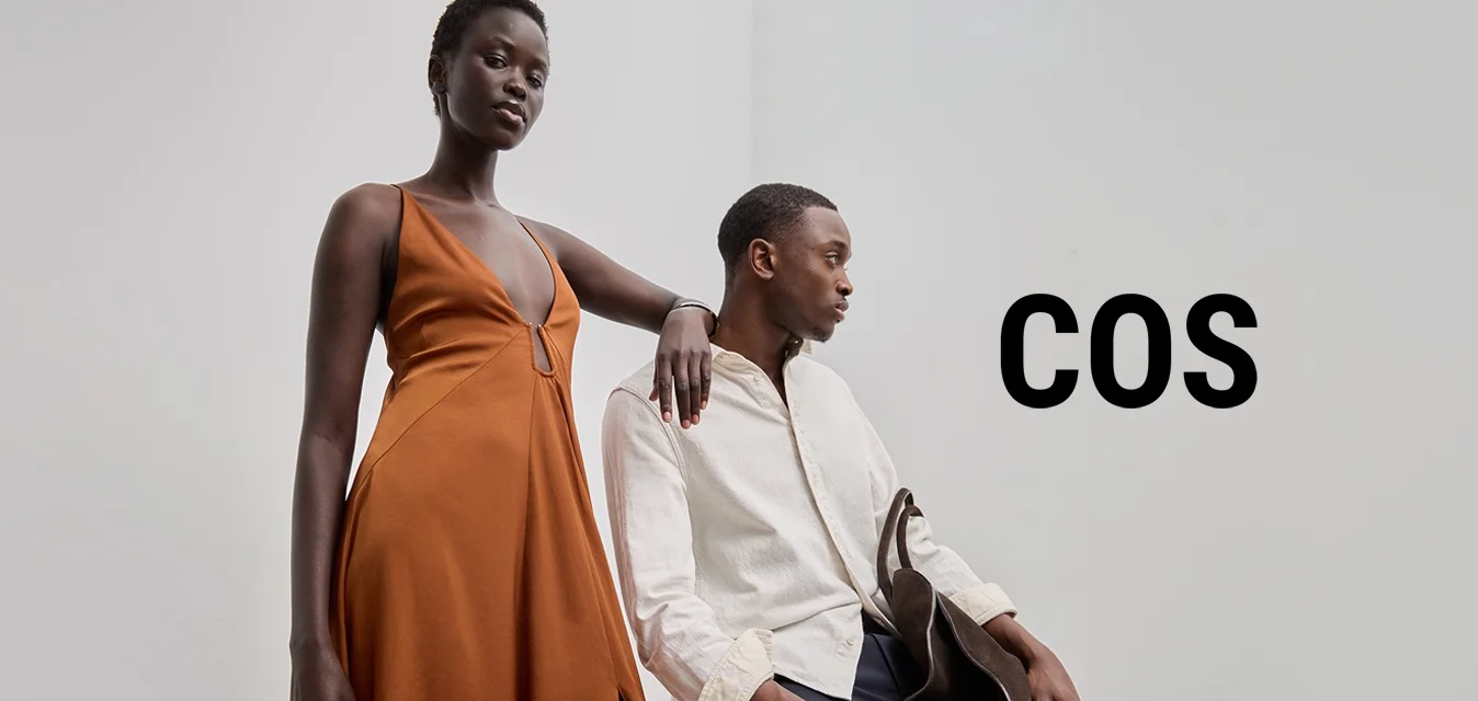 Lungo abito arancione con scollatura profonda e spacco laterale, abbinato a una camicia leggera con texture e pantaloni scuri, impreziosito da una borsa marrone.