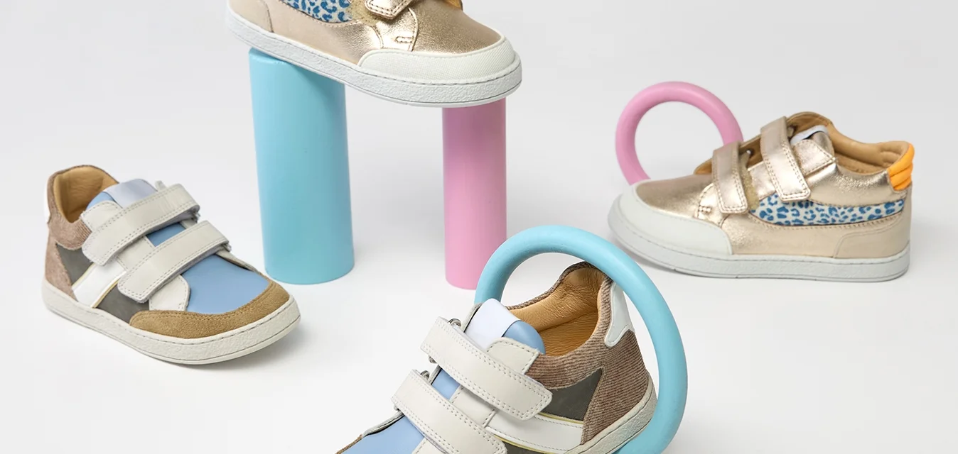 Quattro sneakers colorati per bambini con fasce in velcro esposti su e intorno a forme geometriche pastello blu e rosa su sfondo bianco.