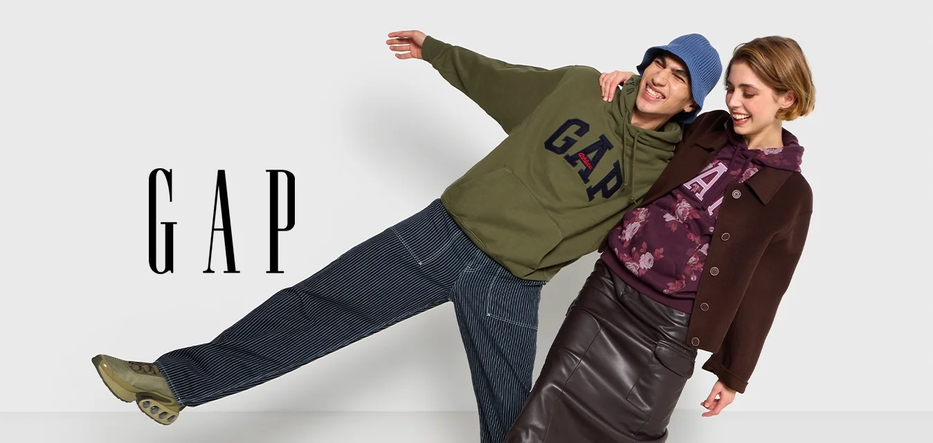 Due modelli si appoggiano in modo giocoso a un muro, indossando abiti casual. Uno indossa una felpa verde oliva e pantaloni larghi, l'altro un cappotto marrone scuro e una gonna.