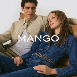 Due persone sono sedute su un divano in pelle marrone chiaro. Una indossa una giacca beige sopra una camicia bianca, l'altra un tuta di jeans blu con una cintura.