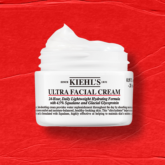 Vasetto bianco di Kiehl’s Ultra Facial Cream aperto, con la crema visibile all’interno, su sfondo rosso testurizzato.