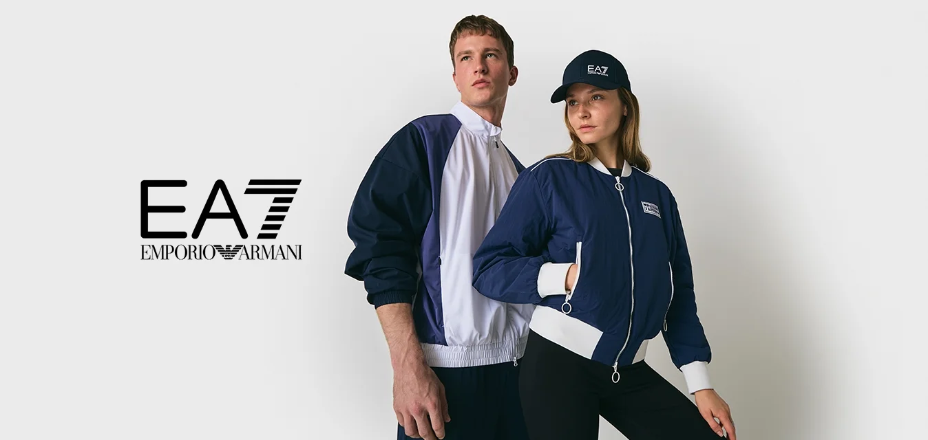 Due giovani adulti in posa con giacche zip-up navy e bianche, uno dei quali indossa un cappellino navy, su uno sfondo chiaro e semplice.