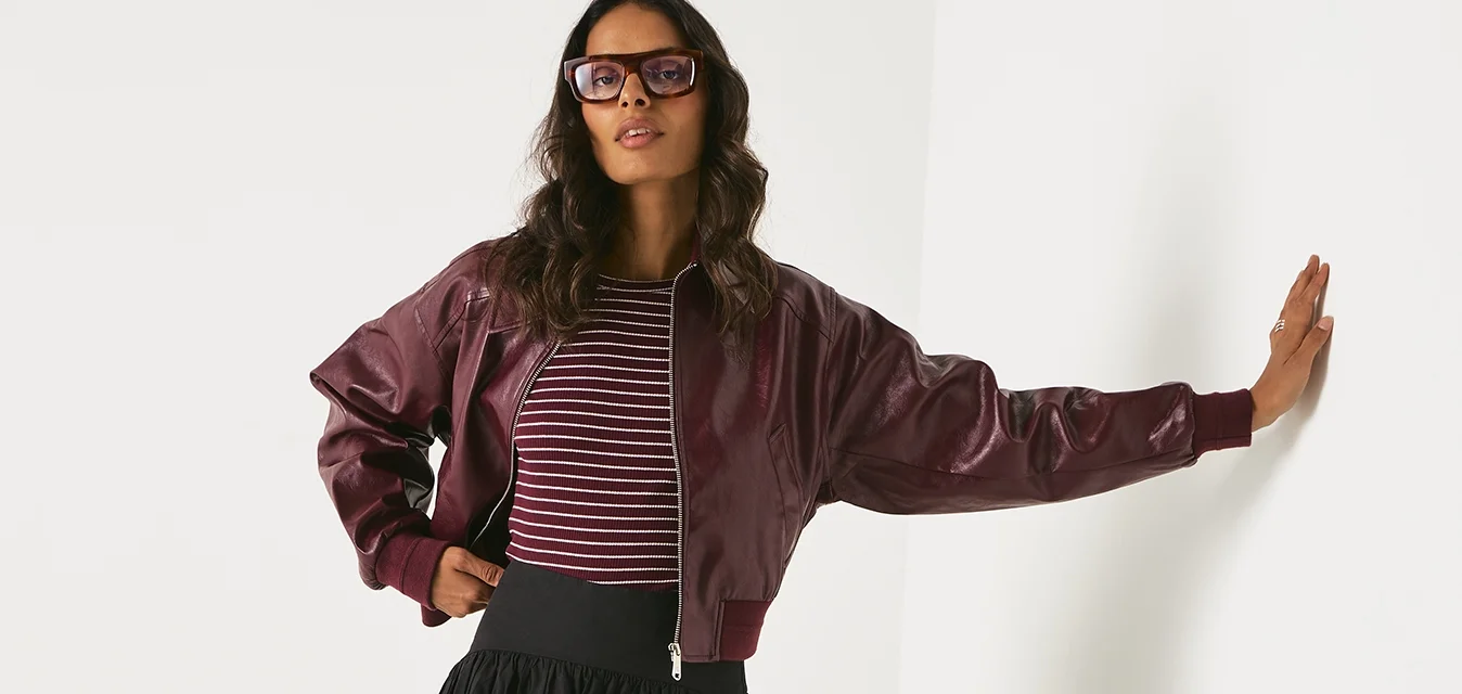 Giubbotto bomber in pelle bordeaux, maglietta a righe, gonna nera plisettata, stivali neri con plateau fino al ginocchio. La modella posa contro un muro bianco.