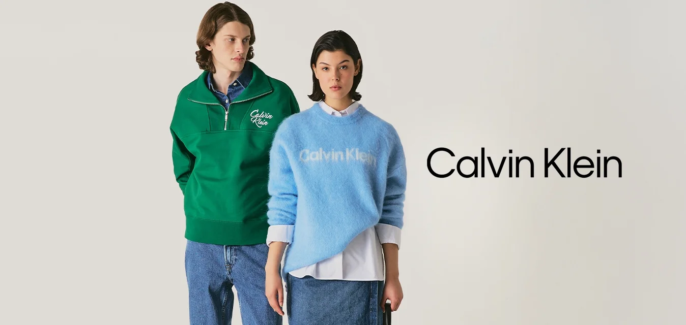 Un uomo in un maglione verde a zip quarti e jeans si trova accanto a una donna in un maglione blu di Calvin Klein, gonna di jeans e che tiene una borsa nera.