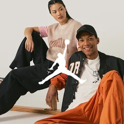 Due persone siedono casualmente su una panchina di legno. L'uomo indossa una giacca arancione e nera, una maglietta bianca, pantaloni arancioni e sneakers grigie. La donna indossa un maglione a maglia rosa e beige con pantaloni neri e sneakers nere.