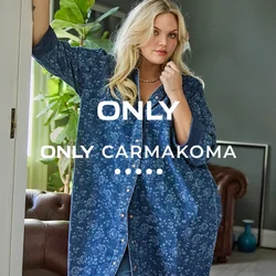 Donna con una maglietta floreale blu larga e jeans appoggiata a un telaio di porta in un soggiorno moderno e luminoso con piante e un divano in pelle.