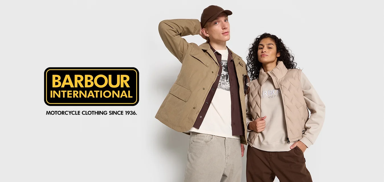 Uomo in giacca beige e cappellino con la mano dietro la testa, donna in gilet beige e felpa con la mano in tasca, entrambi in piedi su uno sfondo bianco.