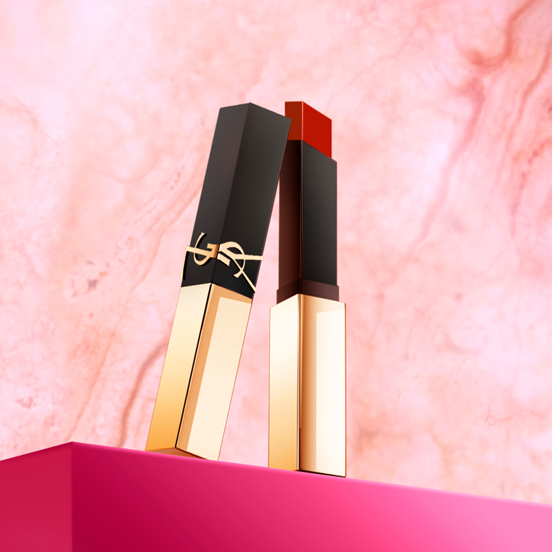 Two Yves Saint Laurent lipsticks, one black and gold, the other gold with a visible red stick, resting on a pink surface against a pink marble wall.