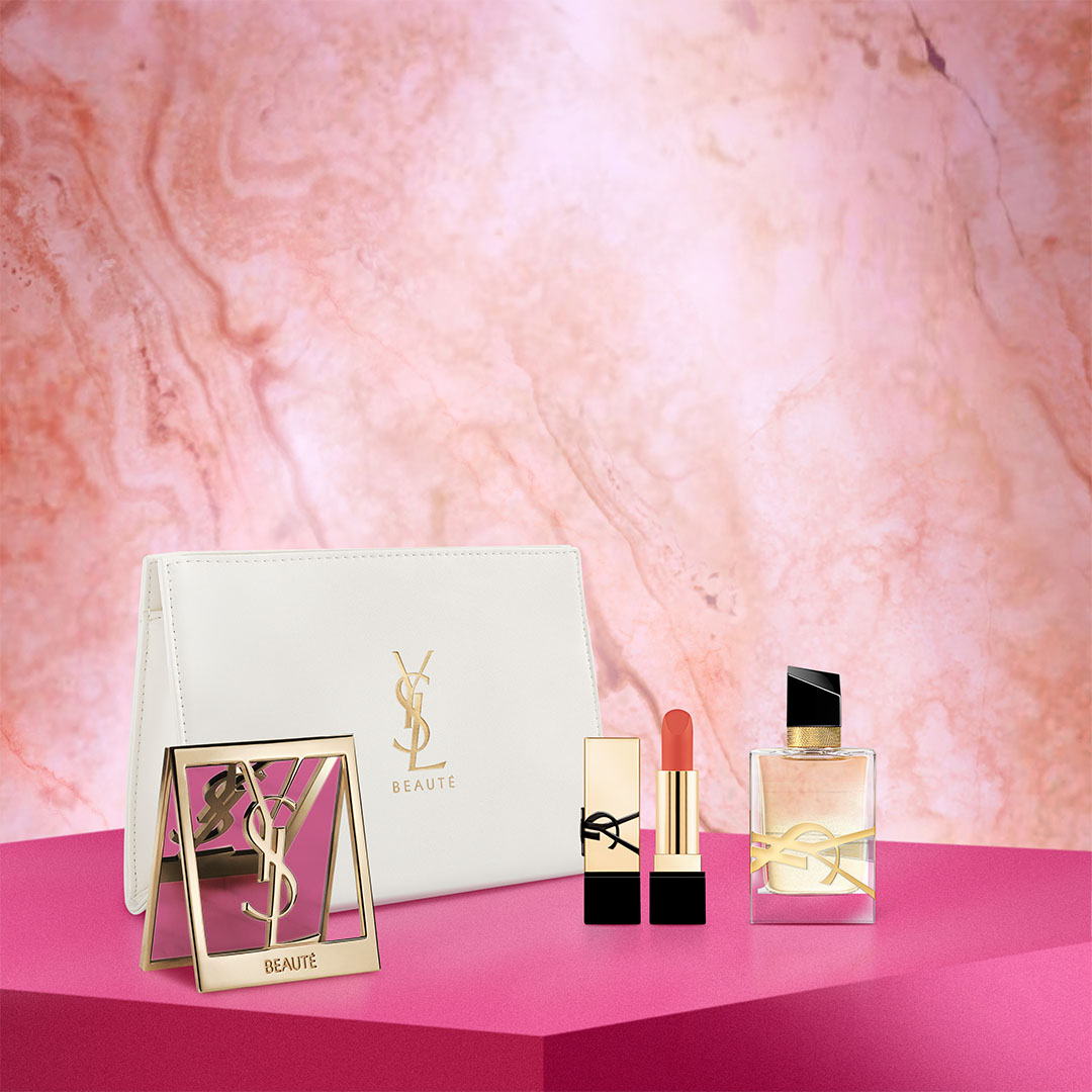 A red Yves Saint Laurent lipstick, a gold bottle of YSL Libre perfume, and a gold compact mirror with the YSL logo and "BEAUTÉ" on it, all displayed on a fuchsia pink surface against a pink marble background.