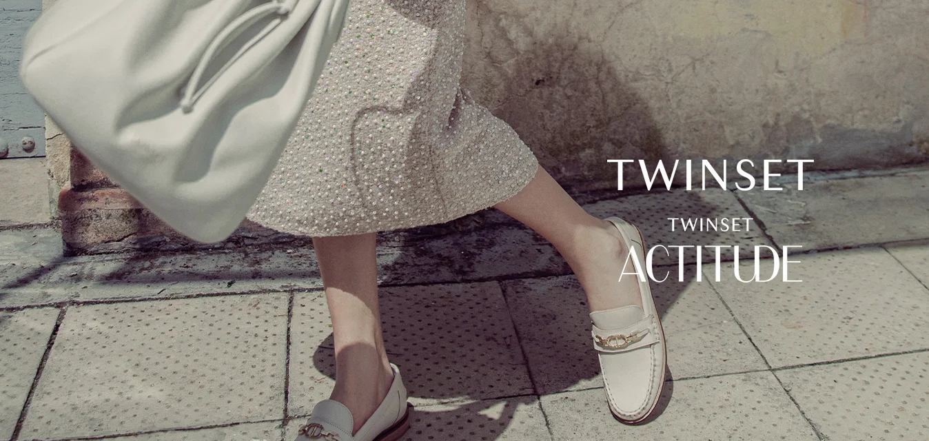 Camicia bianca con maniche ricamate, gonna con fantasia, borsa leggera e mocassini con un dettaglio decorativo. Texture morbide, colori chiari.