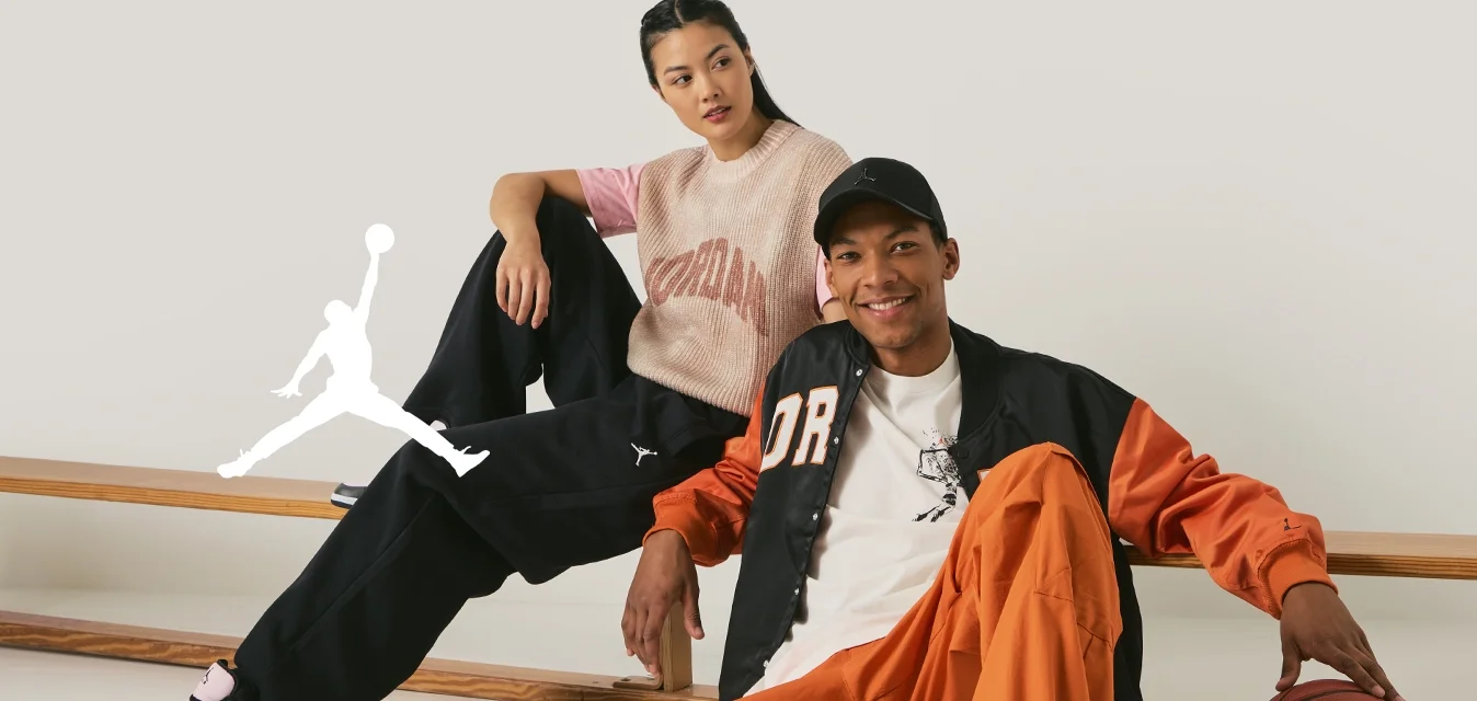 Due persone siedono casualmente su una panchina di legno. L'uomo indossa una giacca arancione e nera, una maglietta bianca, pantaloni arancioni e sneakers grigie. La donna indossa un maglione a maglia rosa e beige con pantaloni neri e sneakers nere.