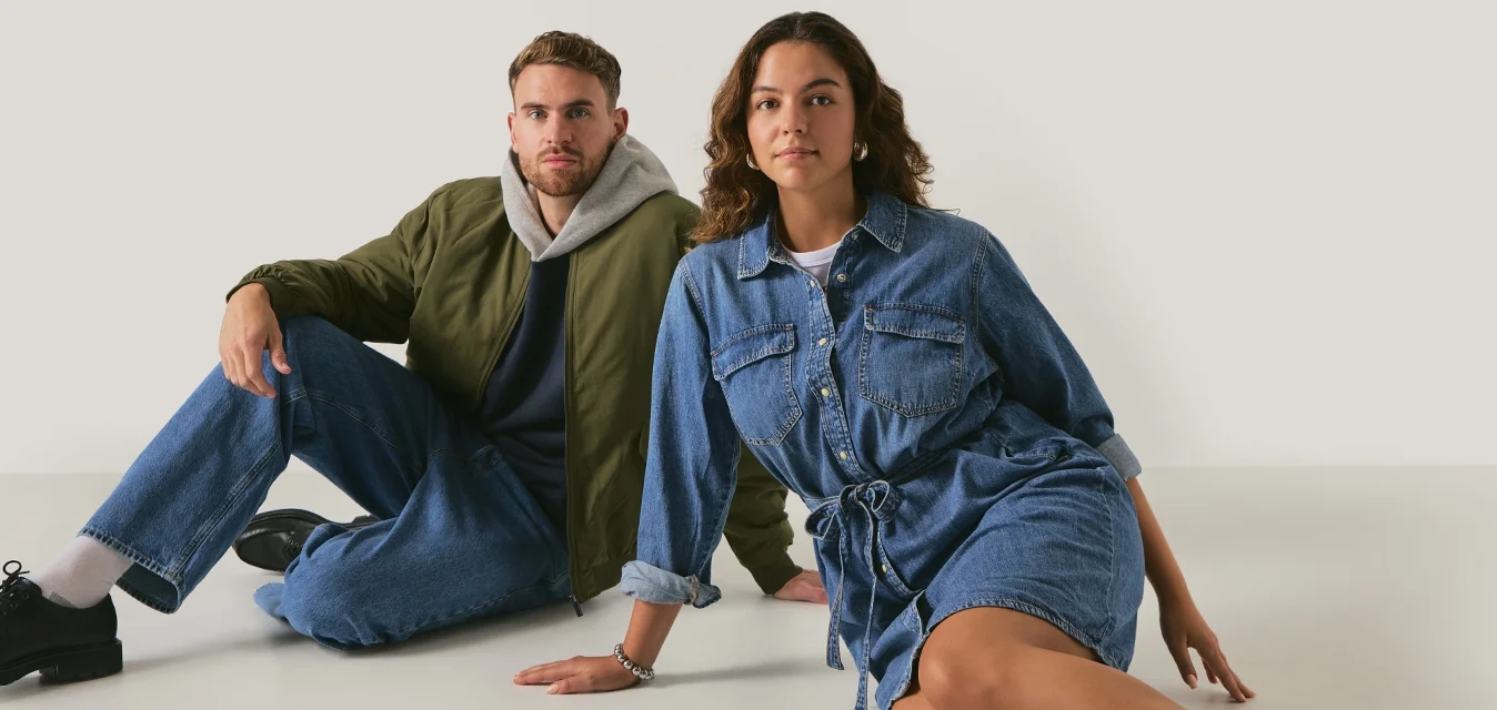 Due modelli seduti su uno sfondo neutro. Il maschio indossa una giacca bomber verde oliva, jeans blu e scarpe nere. La femmina indossa un vestito di denim con cintura annodata in vita e calzini beige con scarpe nere.