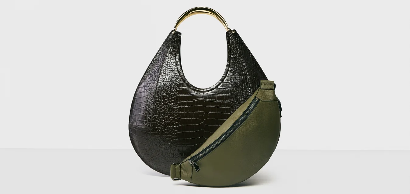 Borsa nera in texture coccodrillo con manico dorato accanto a borsa a tracolla mezzaluna verde oliva con zip nera su sfondo bianco.