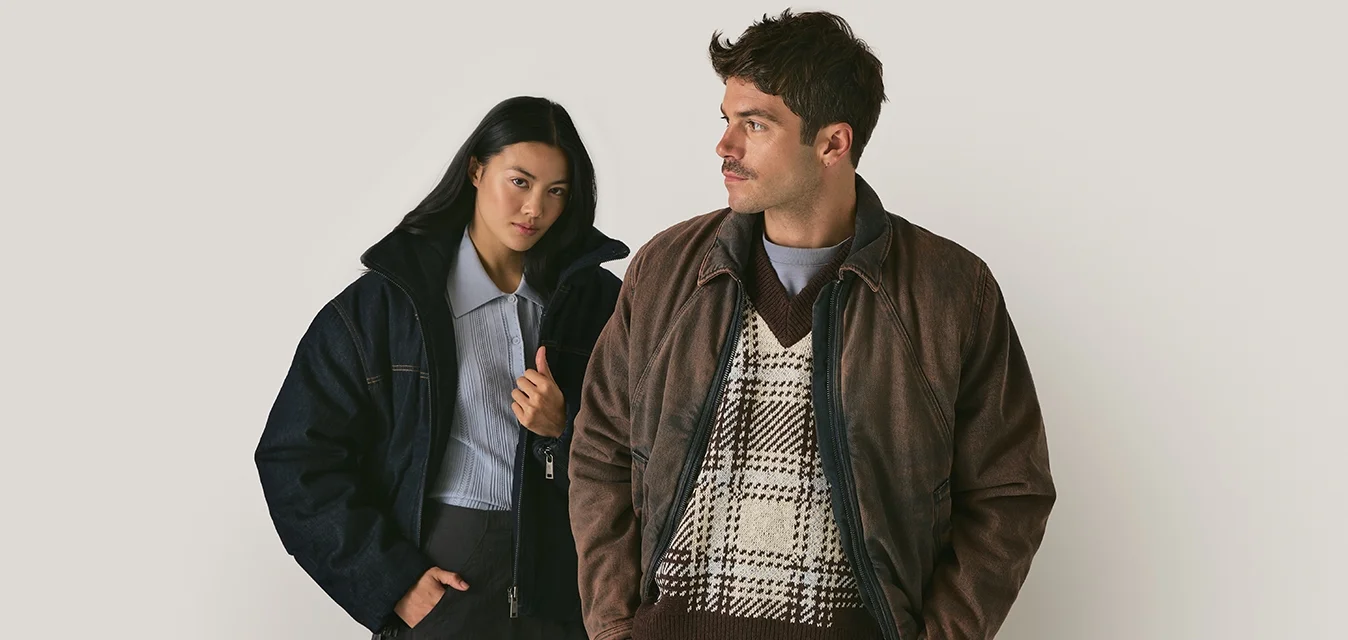 Due persone indossano giacche: una in un piumino in denim scuro e l'altra in una giacca di pelle marrone sopra un maglione a motivo. Entrambi hanno una vestibilità slim.
