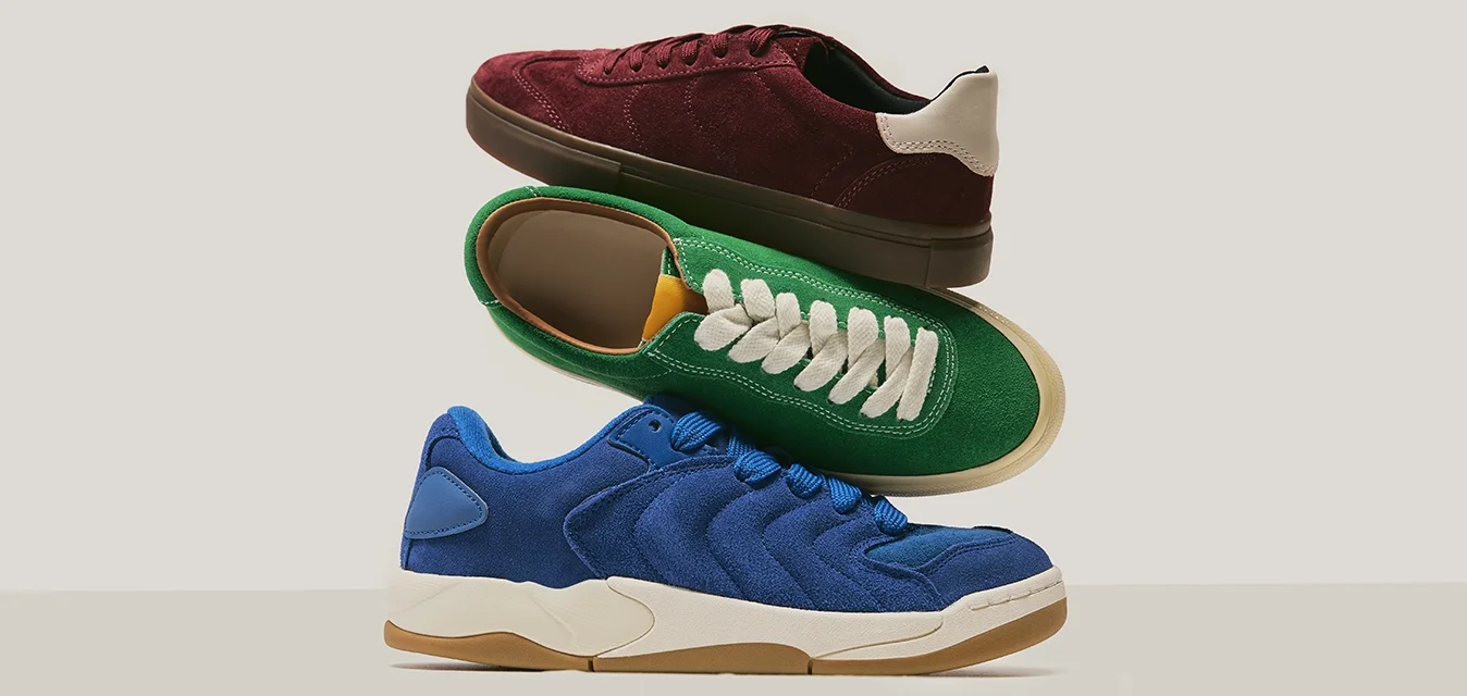 Tre sneakers in suede impilate: una blu, una verde con lacci bianchi e una bordeaux con dettagli beige sul tallone, su uno sfondo neutro.