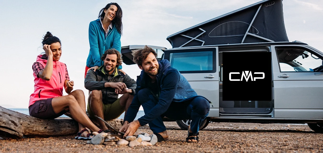 Quattro persone si sono riunite attorno a un piccolo falò su una superficie di ghiaia. Indossano abbigliamento outdoor colorato. Un furgone con il tetto sollevato è parcheggiato nelle vicinanze.